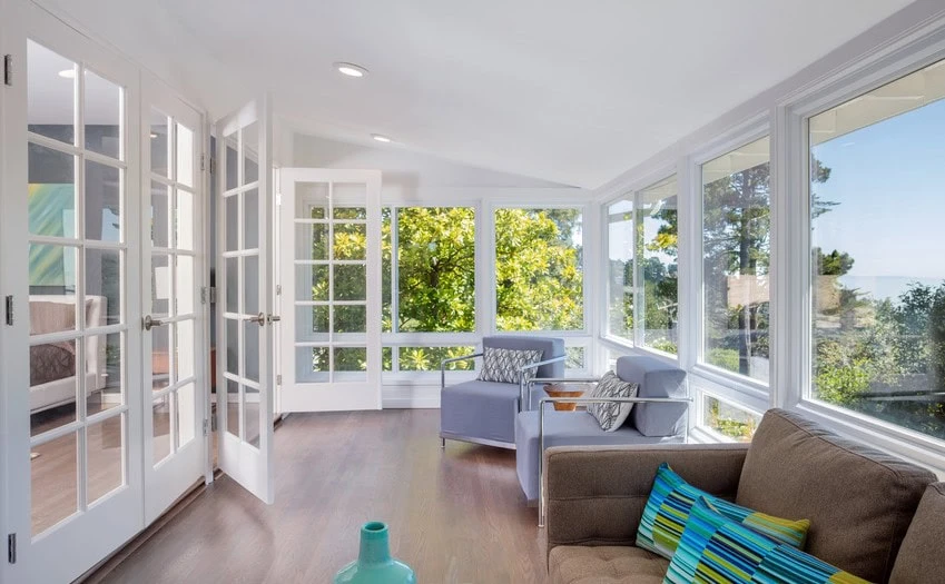 A bright sunroom with large windows and glass doors offers a view of lush greenery outside. The room, nestled in New Castle County, DE, features a brown sofa with colorful pillows, two gray chairs, and light wood floors. A small turquoise vase adds charm to this serene setting.