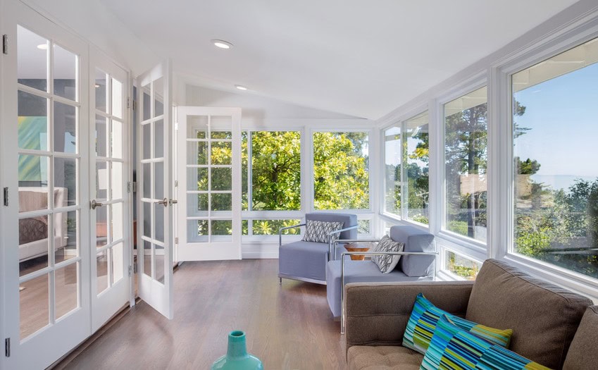 A bright sunroom with large windows and glass doors offers a view of lush greenery outside. The room, nestled in New Castle County, DE, features a brown sofa with colorful pillows, two gray chairs, and light wood floors. A small turquoise vase adds charm to this serene setting.