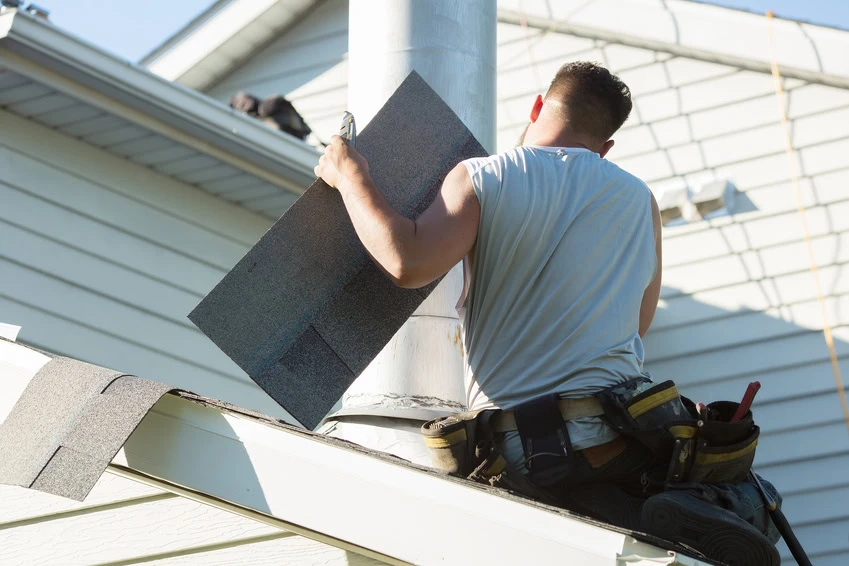 A roofer in a tool belt installs roofing shingles around a chimney on a sloped house roof during daylight.