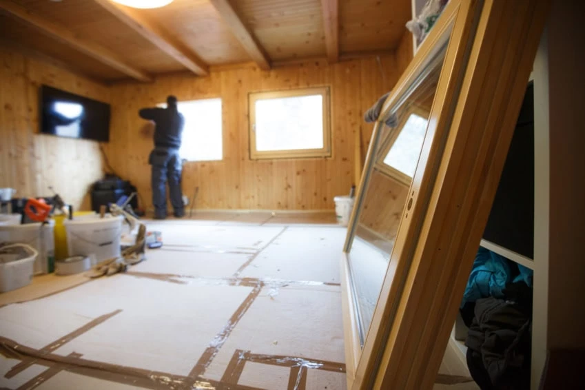 A contractor installs or repairs a window in a wooden room with a slanted ceiling. The floor is covered with protective paper, and tools, buckets, and a large window frame are visible in the foreground.
