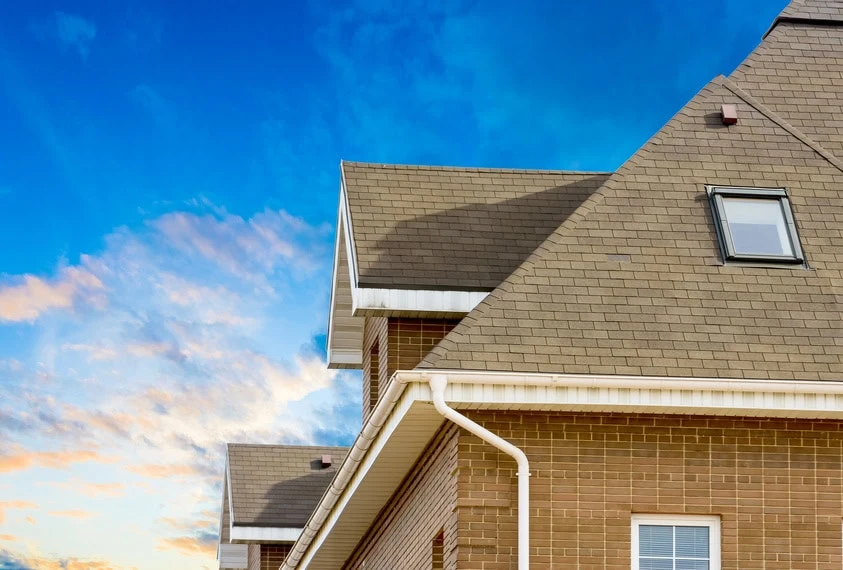Close-up view of a brick house with beige shingle roofing by a trusted roofing contractor, white gutters, and a window on the top floor, set against a bright blue sky with scattered clouds.