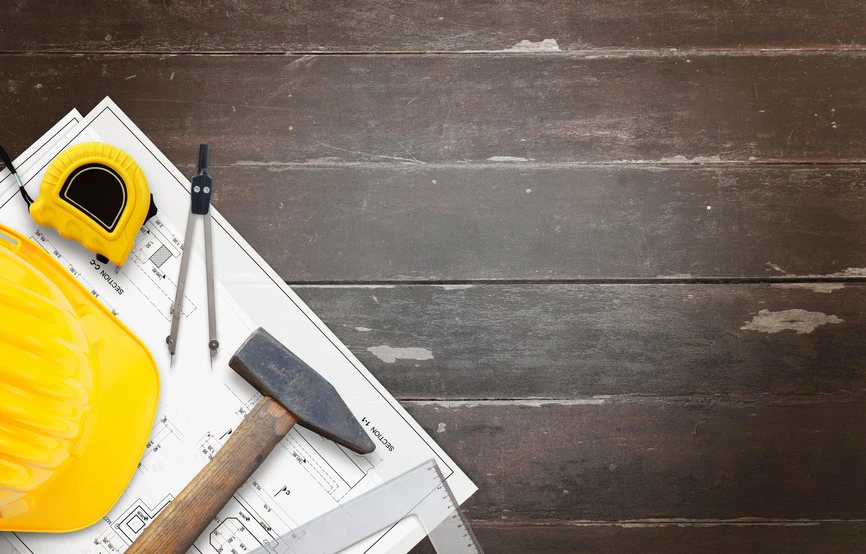 Yellow hard hat, measuring tape, hammer, compass, and ruler on top of architectural blueprints, all resting on a dark wooden surface with empty space on the right.