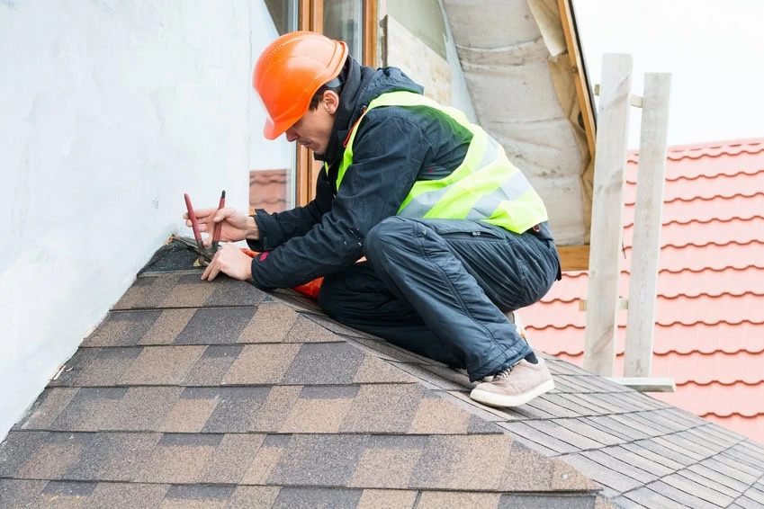 A roofing contractor wearing an orange hard hat, reflective vest, and dark clothing is kneeling on a shingled roof, using a pencil and ruler to mark measurements near the wall of a building.