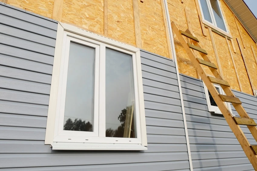 A house exterior partially covered with light gray siding being installed; the upper section shows exposed plywood sheathing. A white-framed window and a wooden ladder leaning against the house are visible.