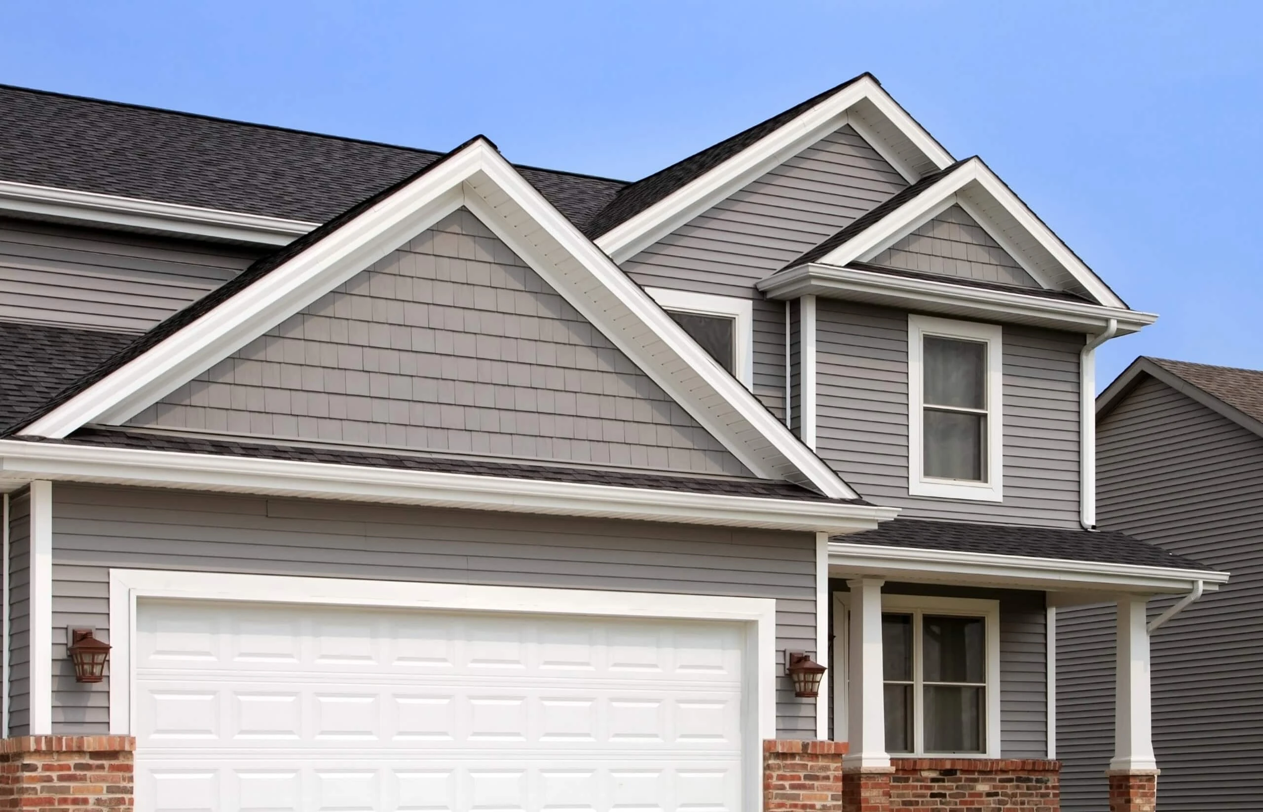 Modern suburban house with gray siding, white trim, and a shingled roof. A large white garage door and a small porch enhance the front. Two windows are visible under the clear blue sky—perfect for those needing roof repair in New Castle County, DE, by an expert roofer.