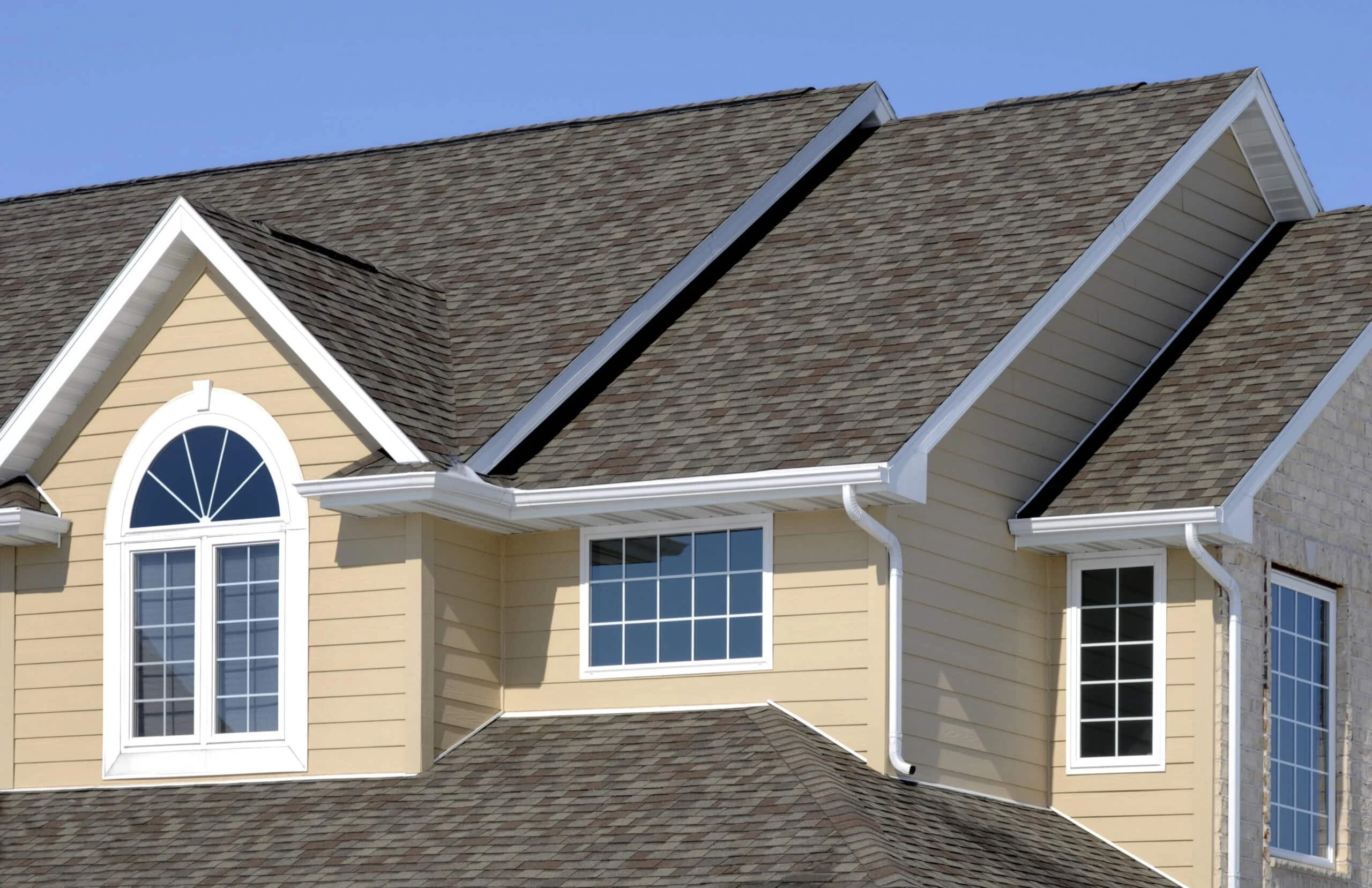 A close-up view of the upper section of a house with beige siding, white trim, multiple windows, and dark gray shingled roofs, showcasing expert roof repair installation against a clear blue sky.