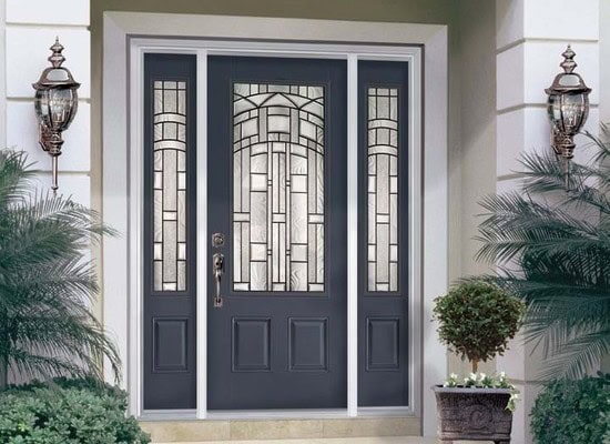 A dark gray front door with decorative glass panels is flanked by two sidelights, framed by light walls and two lantern-style wall sconces. Potted plants enhance the elegant entrance, reflecting the meticulous care common to roof repair in New Castle County, DE.