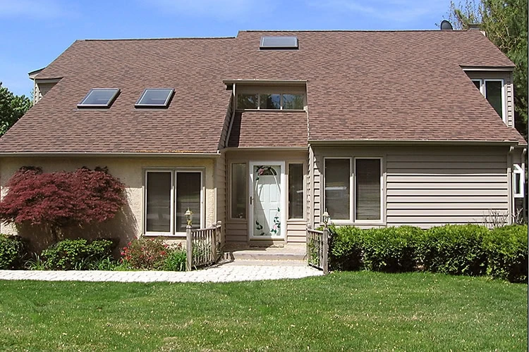 A suburban house with a brown, sloped roof in need of expert roofer services in New Castle County. It features skylights, light-colored siding, a decorative front door, and a path winding through a well-kept lawn and bushes, with a small red tree on the left.
