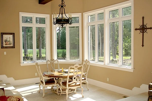 The dining area features a wooden table with six chairs by large windows overlooking a lush DE garden. A decorative chandelier hangs from the ceiling, while a framed picture and cross adorn the beige walls. Natural light fills this serene space, untouched by recent roof repair in New Castle County.