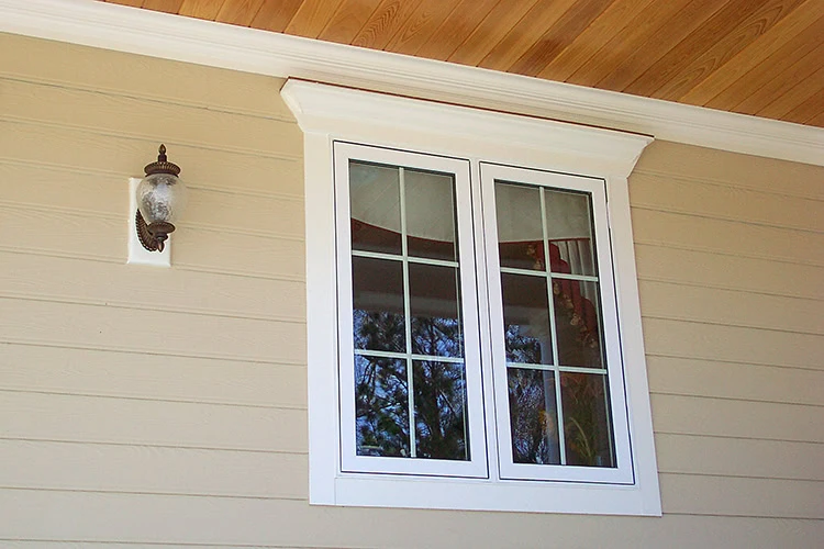 A beige exterior wall with a double glass window framed in white, featuring grid-style panes, reflects the quality craftsmanship of roof repair New Castle County, DE. A decorative lantern with a frosted glass shade is positioned to the left of the window beneath the wooden-finished ceiling.