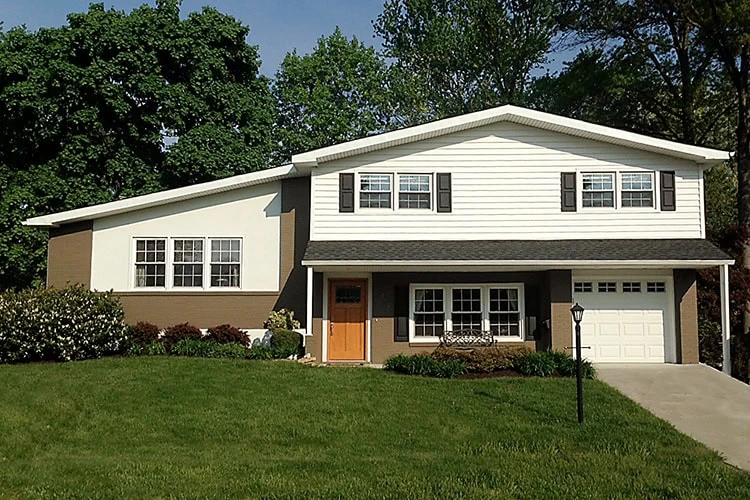 A two-story suburban house with a white and brown exterior stands proudly in New Castle County, DE. Featuring a sloped roof recently inspected by a local roofer, it has multiple windows with shutters, a wooden front door, and is embraced by a single garage and meticulously maintained lawn and trees.