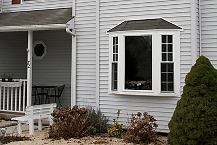 A light gray house exterior with a bay window featuring four glass panels crafted by a skilled roofer in New Castle County. The window is flanked by a small porch with a black table and chairs. In the foreground, there are shrubs and a leafless bush on a stone-covered ground.