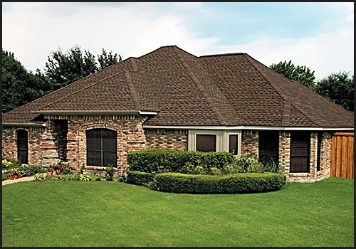 A one-story brick house with a brown, pitched roof showcases the skilled craftsmanship of a roofer from New Castle County, DE. Surrounded by a neatly trimmed lawn and vibrant shrubs and flowers, it features large, dark-framed windows against a backdrop of trees under a clear sky.