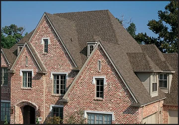 A large brick house with multiple steep gables and gray asphalt shingles recently underwent roof repair in New Castle County. The symmetrically aligned windows on the front facade complement the surrounding greenery and trees beautifully.
