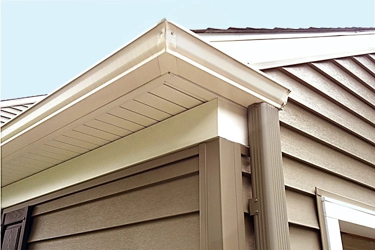 Close-up of a house corner in New Castle County featuring beige vinyl siding, a white overhang, and a downspout. The siding boasts a textured finish, and the overhang displays vented soffit panels. Trustworthy roofer services ensure the horizontal shingles stand firm against the clear blue sky.