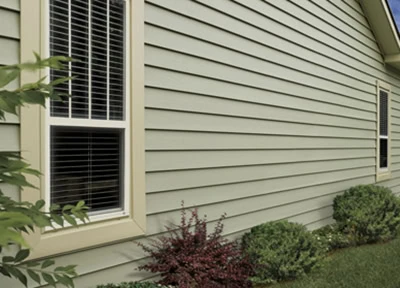 A beige house exterior in New Castle County, adorned with horizontal siding, features two windows with white blinds. Small bushes and a leafy plant grace the front, while a patch of green grass completes the charming scene.