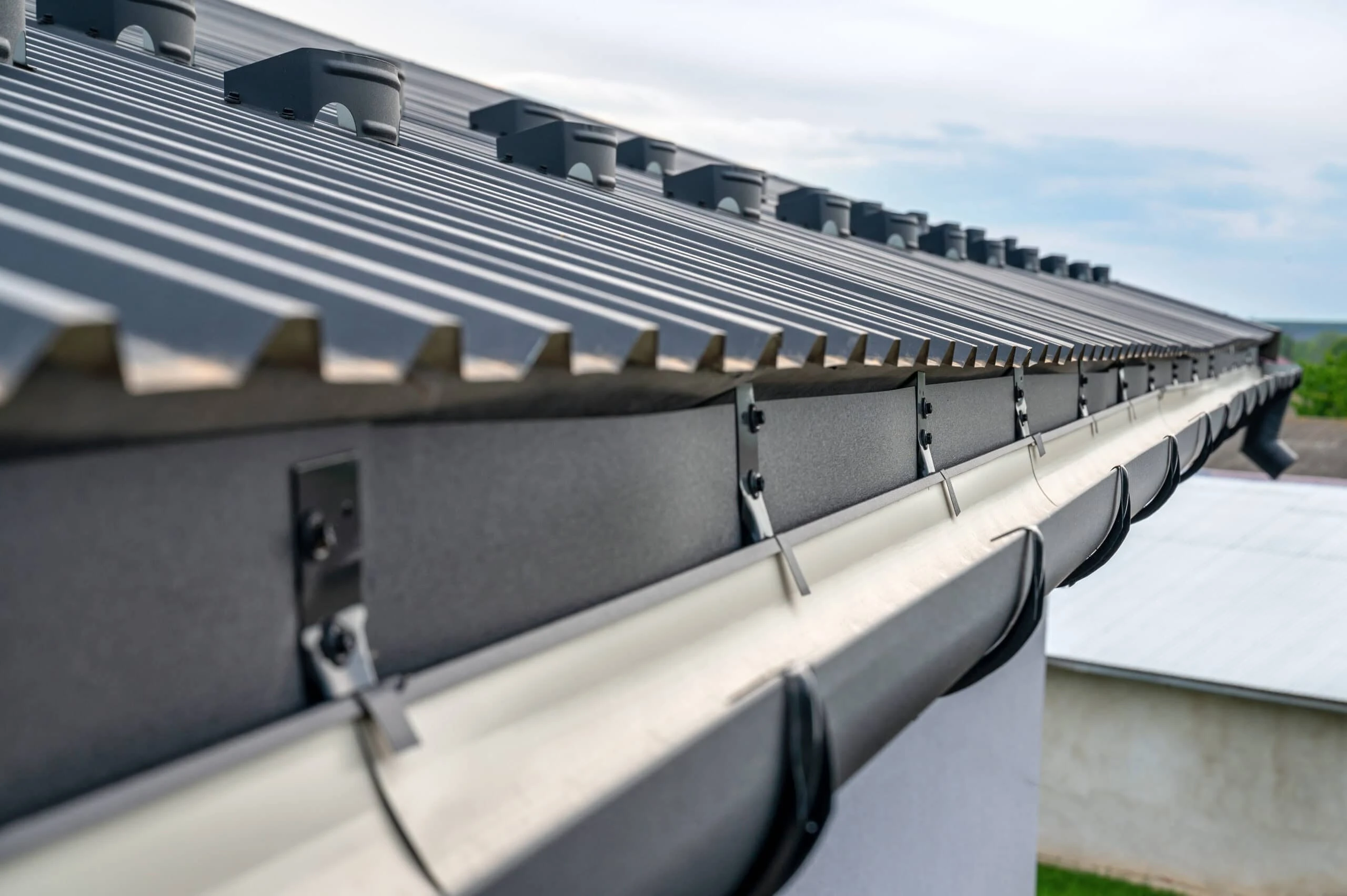 Close-up view of a metal roof in New Castle County, DE, with solar panel mounts along the edge. Below the roof, a white gutter system is attached, curving along the side of a building. The sky is partly cloudy in the background. Perfect for those considering roof repair.