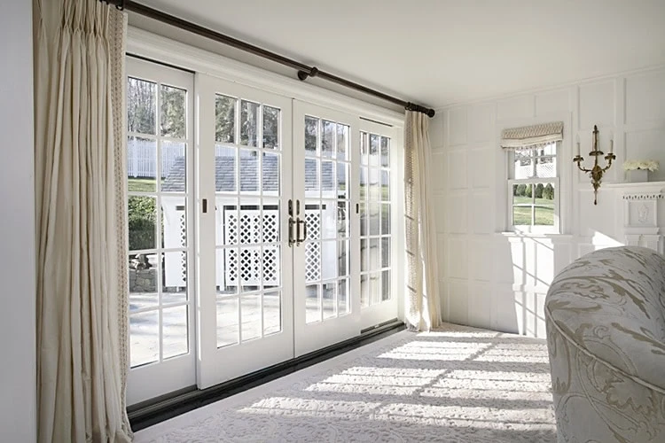 Bright living room with large glass sliding doors leading to a patio, perfect for enjoying the New Castle County sunshine. The room features white walls, a patterned rug, a sofa, and a classic light fixture. Sunlight casts shadows through the doors framed by cream-colored curtains.