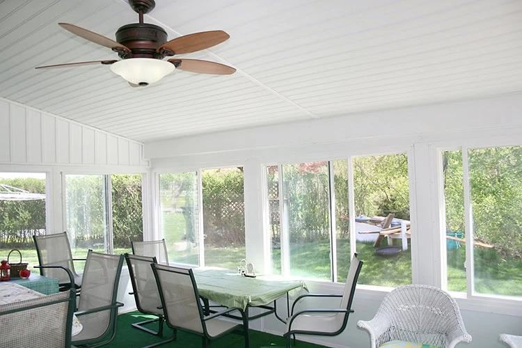 Bright sunroom with white walls and ceiling, featuring a roofer-approved ceiling fan with wooden blades. Large windows offer a view of the lush New Castle County greenery. The room is furnished with a green tablecloth on the table and several wicker and metal chairs.