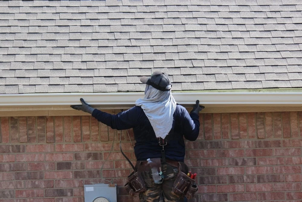 A roofer in New Castle County, DE, wearing a cap and protective gear, stands on a ladder repairing a gutter on a brick house. Gloved hands align the gutter beneath the roof shingles while various tools are neatly tucked in their tool belt.