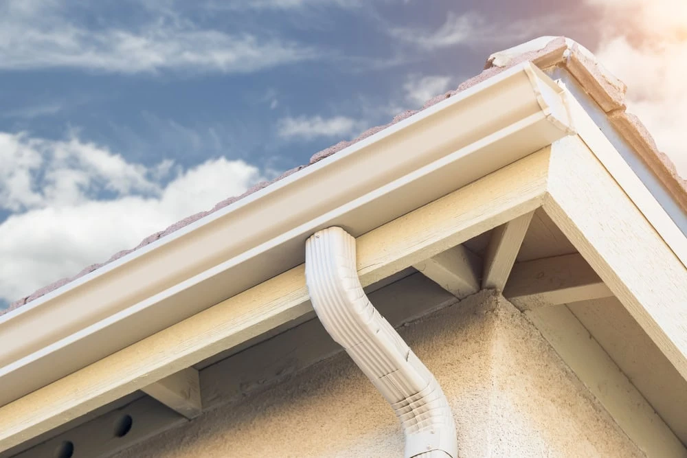 Close-up view of a house's roof corner with a white gutter and downspout under a blue sky with clouds. The light-colored shingles gleam in the sunlight, highlighting the structure's details—a perfect day to consider roof repair in New Castle County, DE.