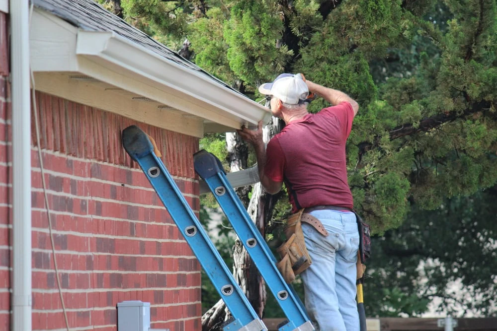 A person in a red shirt and cap stands on a blue ladder, expertly fixing a gutter on a brick house. With a tool belt around their waist and a tree in the background, this New Castle County roofer ensures top-notch repairs.