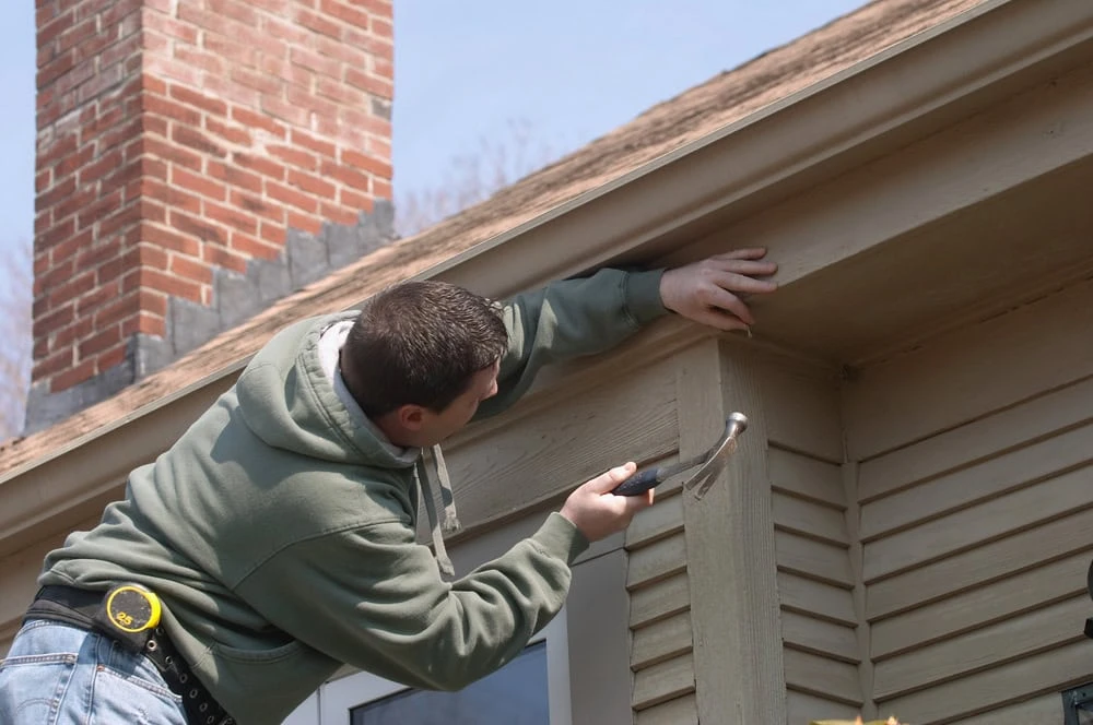 A roofer clad in a green hoodie deftly uses a hammer to repair a wooden section of a house's exterior near the roof. Standing confidently on a ladder with a tape measure clipped to their jeans, they work skillfully under the watchful gaze of New Castle County's iconic brick chimneys.