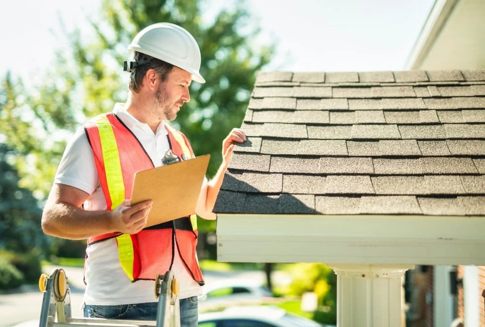 A roofer in New Castle County, DE, wearing a white hard hat and neon safety vest, inspects roof shingles. He holds a clipboard while standing on a ladder next to the house. Trees and a clear sky are visible in the background.