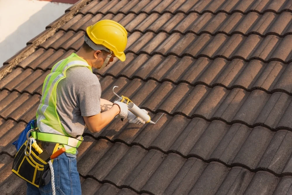 A construction worker in a yellow hard hat and reflective vest uses a caulking gun to seal gaps on a tile roof, providing quality roof repair in New Castle County, DE. He's carrying tools in a belt and is wearing jeans, focused on his work under natural daylight.