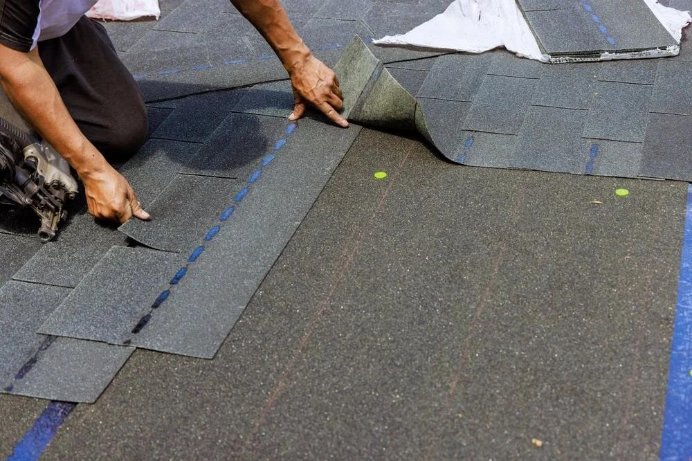 A skilled roofer in New Castle County is laying dark asphalt shingles on a roof. With precision, they use a nail gun to secure each shingle while their hands meticulously adjust the placement. The textured shingles align perfectly with the blue guidelines on the roof.