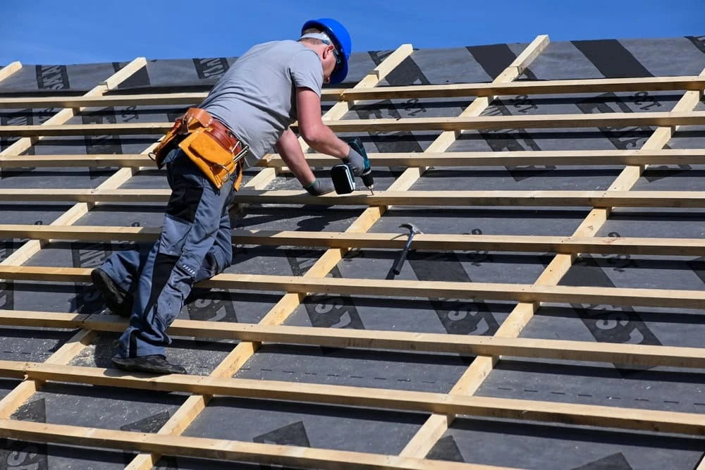 A skilled roofer in New Castle County, wearing a hard hat and tool belt, diligently works on a sloped roof under the clear blue sky. Dressed in a gray shirt and dark pants, they use a power drill to secure beams, showcasing their expertise in roof repair.