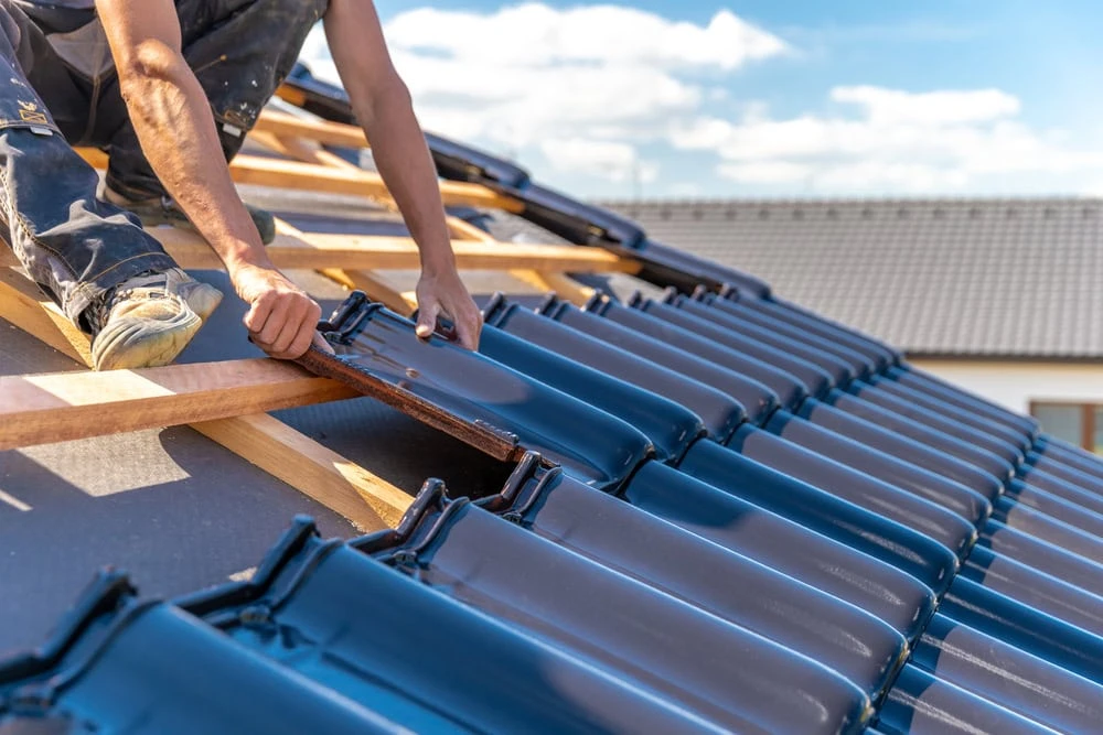 A person is installing dark blue roof tiles on a wooden frame of a house under a partly cloudy sky in New Castle County, DE. The person is wearing work pants, and another roof with grey tiles is visible in the background.