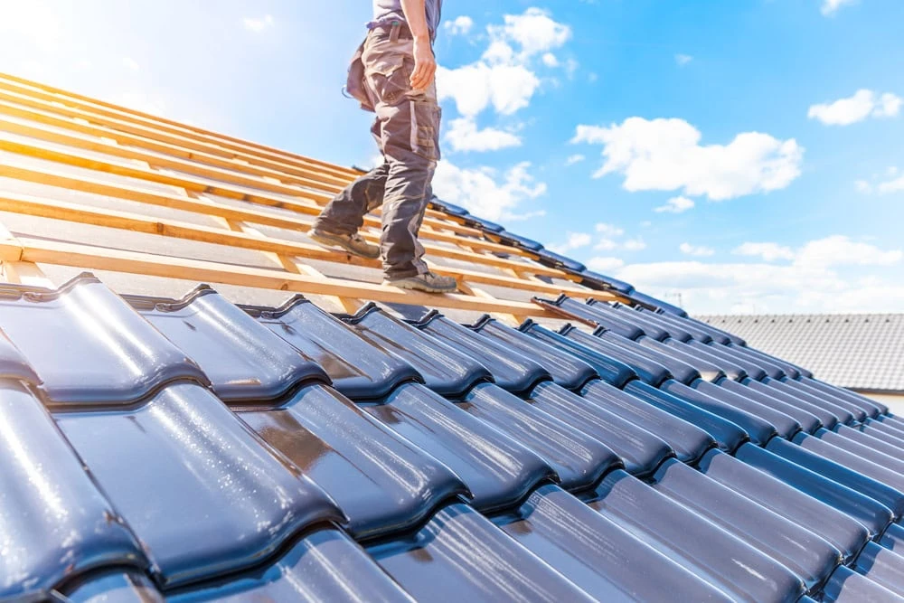 A person stands on a roof under construction in New Castle County, clad in workwear. One side of the roof boasts shiny blue tiles, while wooden beams await completion on the other. Overhead, a bright sky with scattered clouds stretches across the scene.