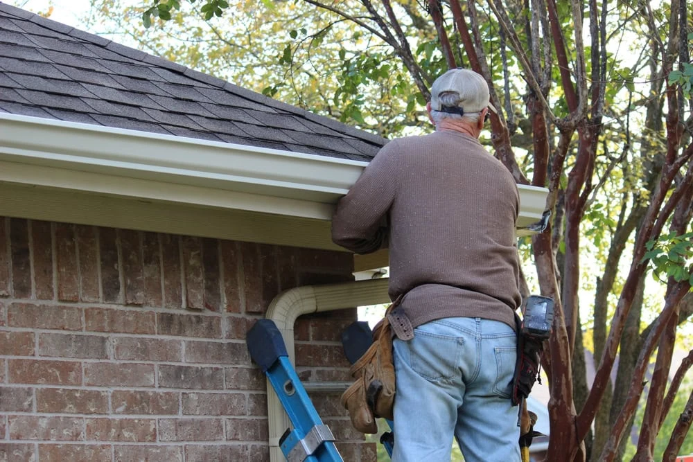 A roofer in a cap and brown sweater is perched on a ladder beside a brick building in New Castle County, DE, diligently working on a gutter. Equipped with a tool belt around their waist, they are framed by the silhouette of a tree in the background.