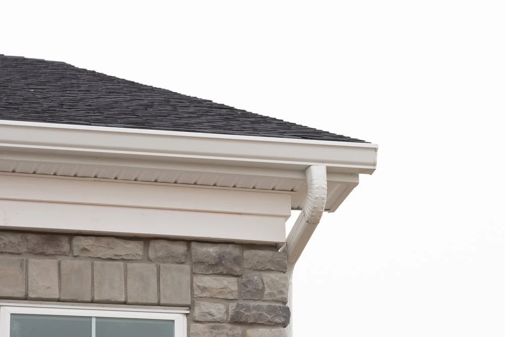 Close-up of a house corner reveals dark shingles expertly laid by a roofer in New Castle County, complemented by a white gutter and beige stone exterior walls. The downspout seamlessly connects to the gutter, framed against an overcast sky.