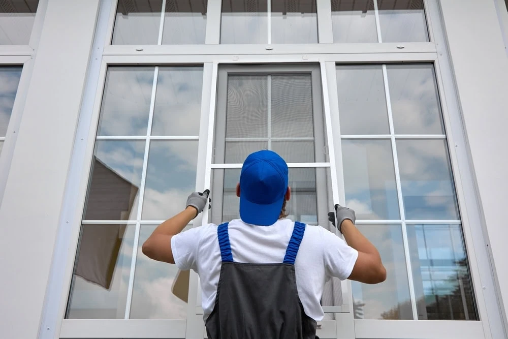 A worker in a blue cap and gloves inspects a large window with multiple panes against a white frame. While assessing potential roof repair needs in New Castle County, DE, the clear glass reflects an overcast sky as the person is seen from behind, intently focused on their task.