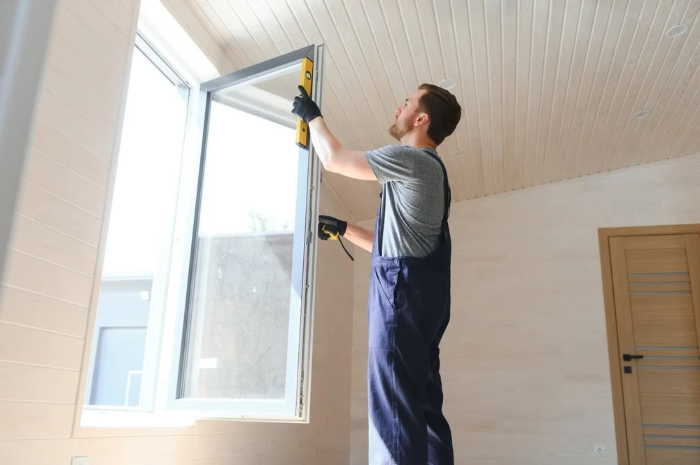 A man in overalls and gloves is expertly installing a window in a bright room with wooden walls and ceiling. Using a level tool for perfect alignment, this roofer from New Castle County, DE, ensures precision as natural light streams through the room's closed wooden door.