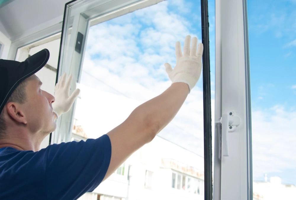 A person wearing a cap and white gloves, perhaps a roofer from New Castle County, installs a large window pane, aligning it into the frame. The bright blue sky with some clouds is visible outside, illuminating the room beautifully with natural light.