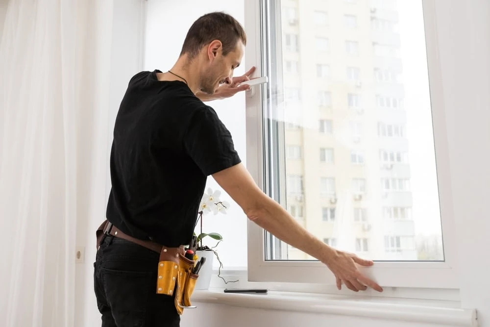 A man in a black shirt and tool belt is expertly installing or adjusting a white-framed window in a bright room. A white orchid graces the windowsill, offering a serene contrast to the bustling view of New Castle County, DE's tall buildings visible through the glass.