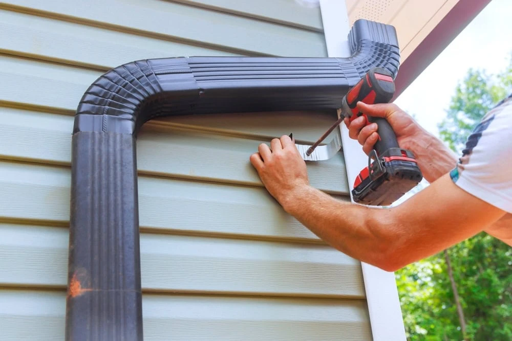 A person uses a red and black power drill to secure a black gutter downspout to the exterior wall of a house in New Castle County, DE. The light blue siding contrasts with the lush trees visible in the background, framing this scene of routine roof repair.