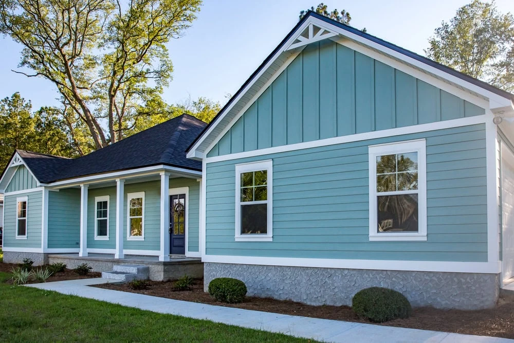 A light blue, single-story house with a dark roof awaits roof repair in New Castle County, DE. It features white trim, multiple windows, and a charming front porch supported by white columns. Surrounded by a lush lawn and trees, the scene is bright under a clear sunny sky.