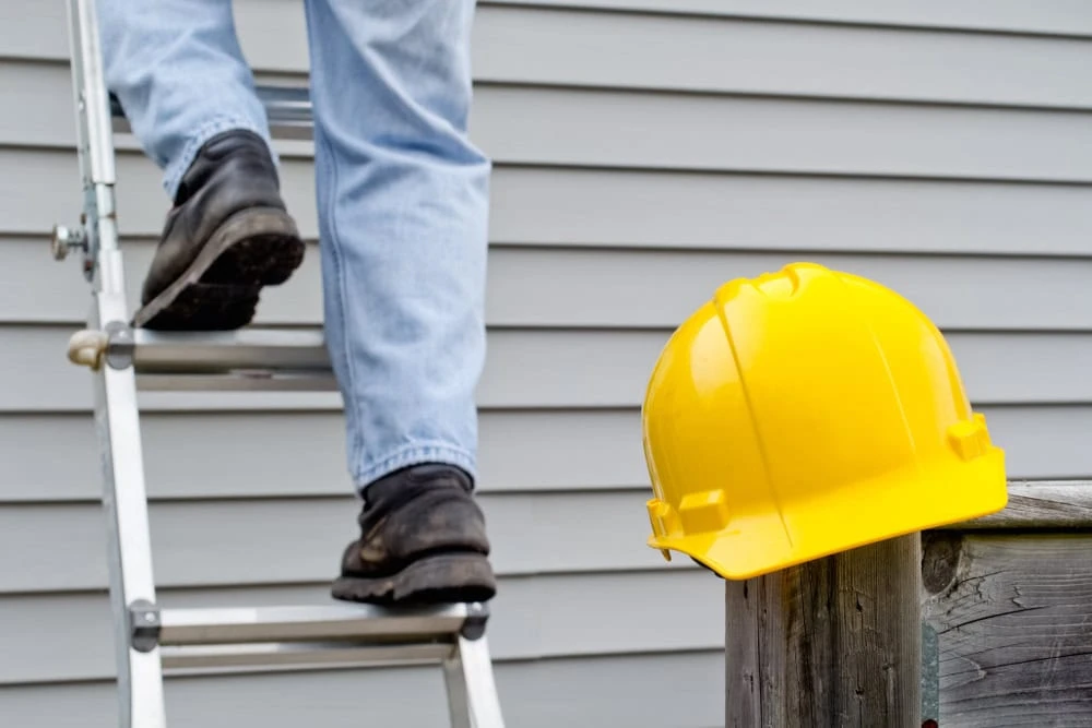 A person wearing jeans and black boots climbs a ladder against a grey siding wall, possibly for roof repair in New Castle County, DE. A yellow hard hat rests on a wooden railing nearby.