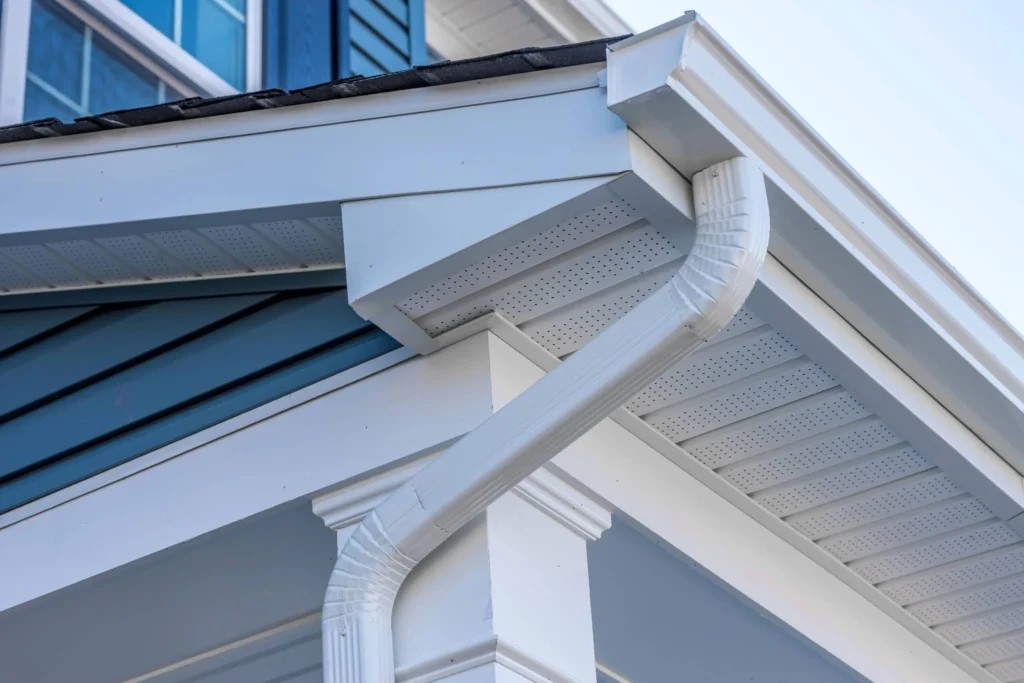 Close-up view of a house corner showing white gutters, downspout, soffit, and roofline against blue siding. The image highlights the clean lines and details of the building’s exterior.
