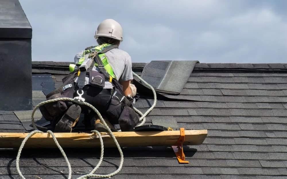 A roofer in New Castle County, DE, wearing a helmet, safety harness, and gear, kneels on a shingle roof to perform repairs. Surrounded by ropes and equipment under a cloudy sky, the construction worker is focused on ensuring the roof is secure and weatherproof.
