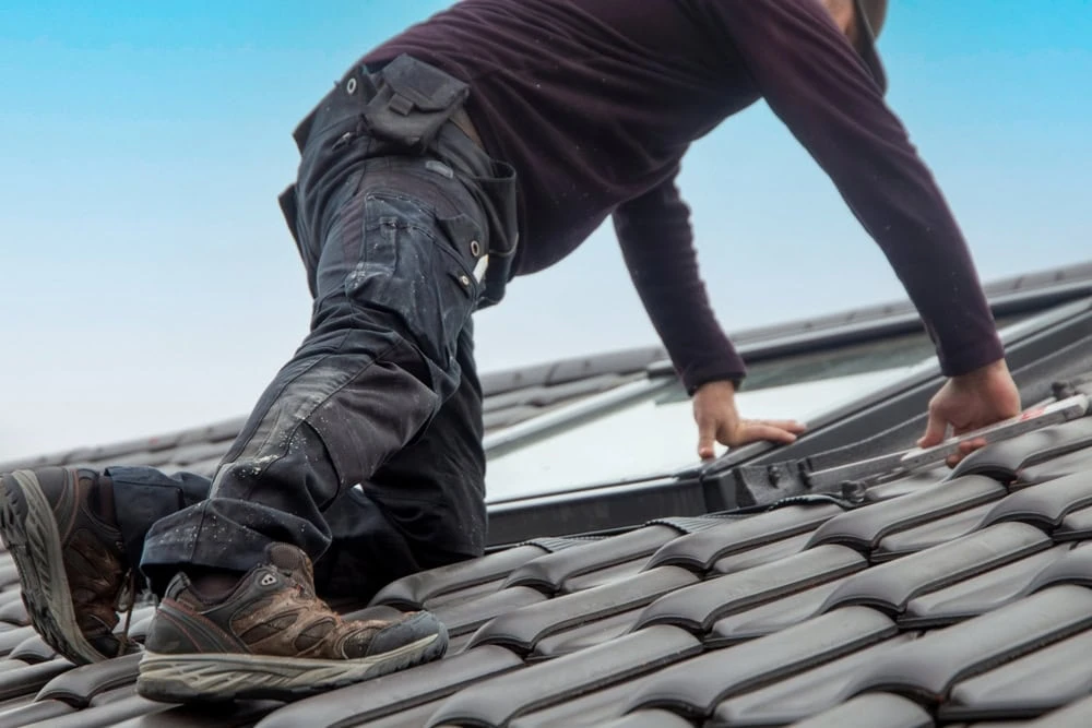 A roofer in dark clothing and hiking boots is kneeling on a textured, brown-tiled roof in New Castle County, DE, adjusting a skylight window. The sky in the background is clear and blue.