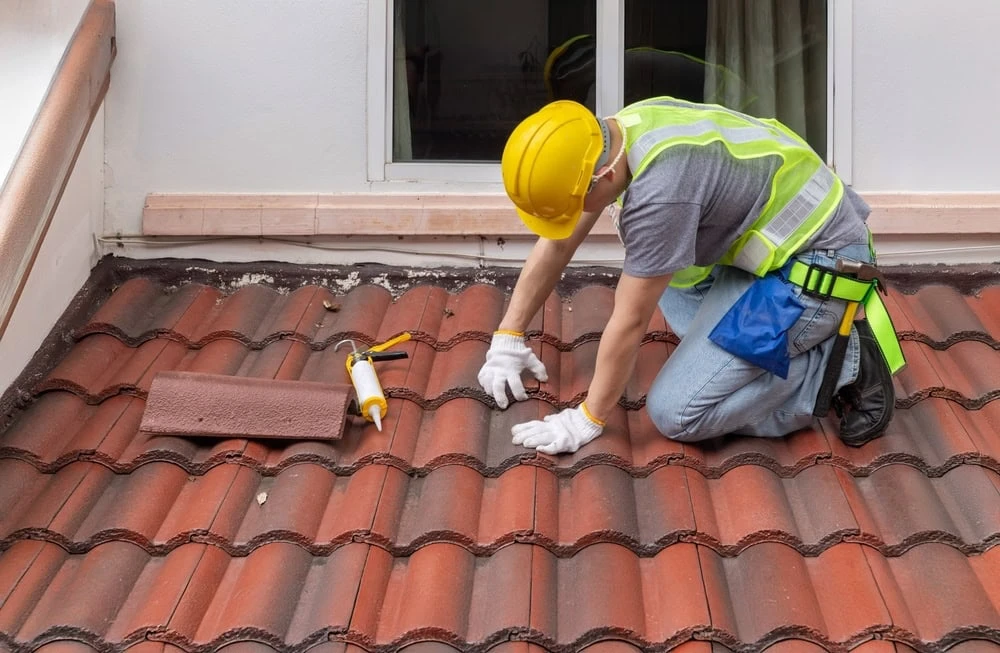 A roofer in New Castle County, dressed in a yellow hard hat and safety vest, kneels on a red tiled roof. They expertly apply sealant to a tile with a caulking gun, tools neatly arranged in their utility belt and another tile ready at hand.