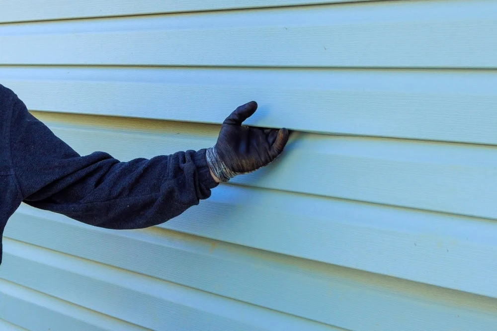 A person wearing a dark glove and sweater is touching light blue vinyl siding on a building. The image highlights the hand and arm, capturing the texture of the siding panels, frequently encountered by roofers in New Castle County during roof repairs.
