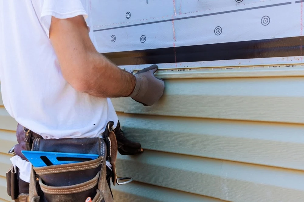 A roofer in New Castle County, DE, is installing light green vinyl siding on a house. Wearing a white t-shirt and tool belt stocked with tools, the focus remains on the precise craftsmanship evident in the installation process and the person’s skilled hands and torso.
