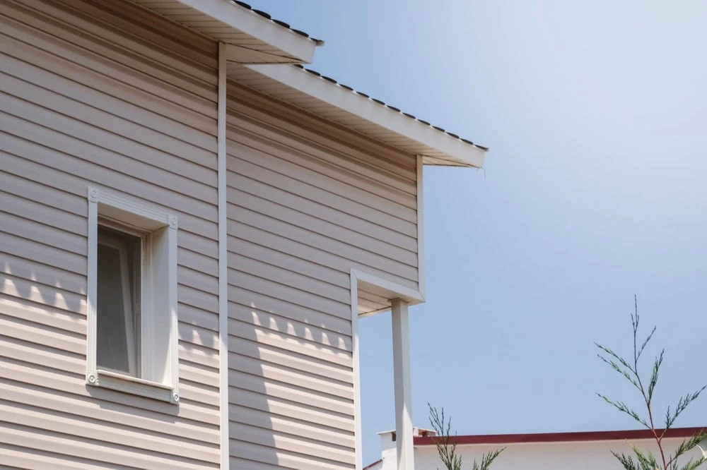 A close-up view of a modern house's exterior with beige vinyl siding and a white-framed window. The roof edge showcases the fine work of a roofer from New Castle County, DE, set against a clear blue sky, while greenery peeks through in the foreground.