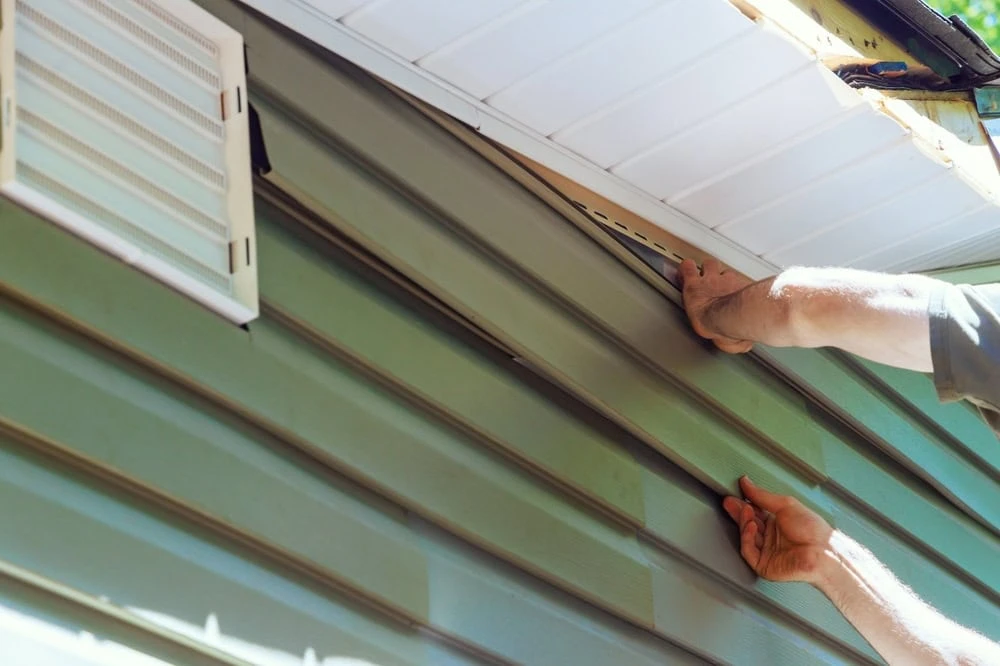 Close-up of a person’s hands installing green vinyl siding on the exterior of a house, under the eaves, with a vent visible on the wall nearby.
